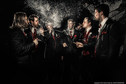 The Finer Things — Probably my 2nd-favorite image of all time came from Matt and Lindsey’s wedding reception held at Nestleton Waters Inn: the groomsmen enjoying a quiet moment with fine cigars. While it looks like dusk, we actually used High Speed Sync (HSS) to overpower the daylight and create this moody, cinematic atmosphere. This shot was a true team effort — my partner Teresa was instrumental, perfectly positioning the rear strobe to catch the smoke without shadows and provide that critical separation from the background. We used a remote two-light setup (rear and camera-left) to turn a daytime scene into a high-drama portrait. Teresa’s job was the most important one here. In photography, smoke is almost invisible unless it is backlit.  If you look closely you can see her hiding behind the groomsmen in this behind-the-scenes snapshot, taken at almost the same moment by the resort owner standing beside me.